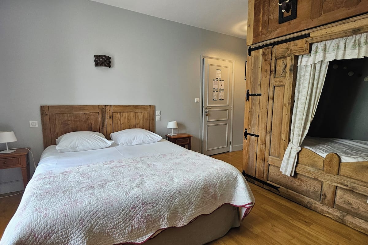 Een slaapkamer met een houten bed, nachtkastje, een kledingkast met gordijn en een kleine wandversiering.