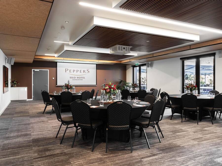 A conference room with a large screen displaying 'PEPPERS SILO HOTEL LAUNCESTON,' a table set with chairs, and a view of the outdoors through large windows.