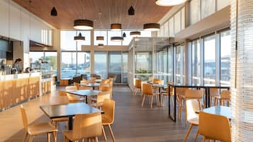 A modern cafe with wooden flooring, tables, chairs, and a counter with a person working. Large windows provide views of the city.