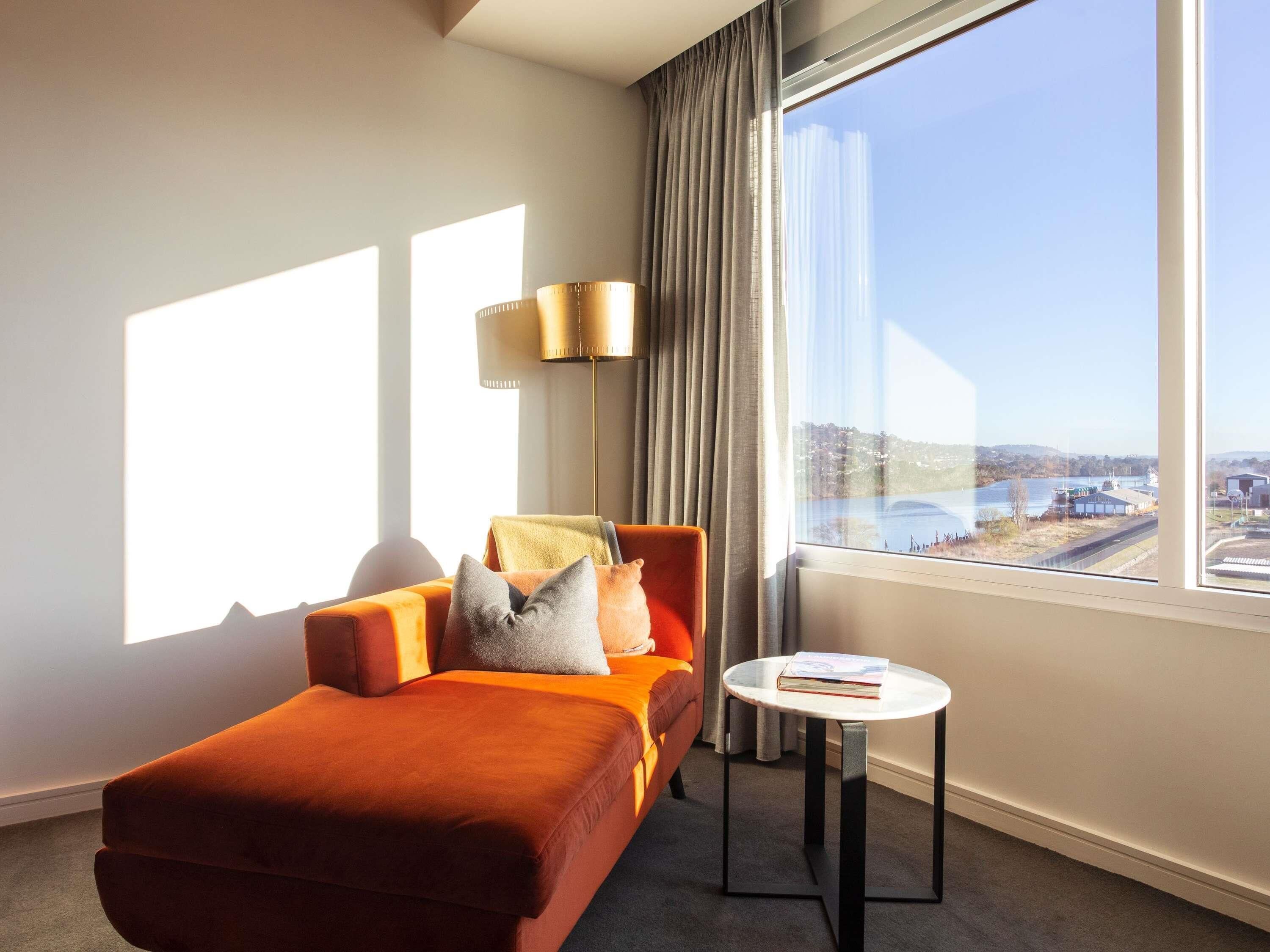Una habitación de hotel moderna con un ventanal grande, un sofá, una mesita y una lámpara.