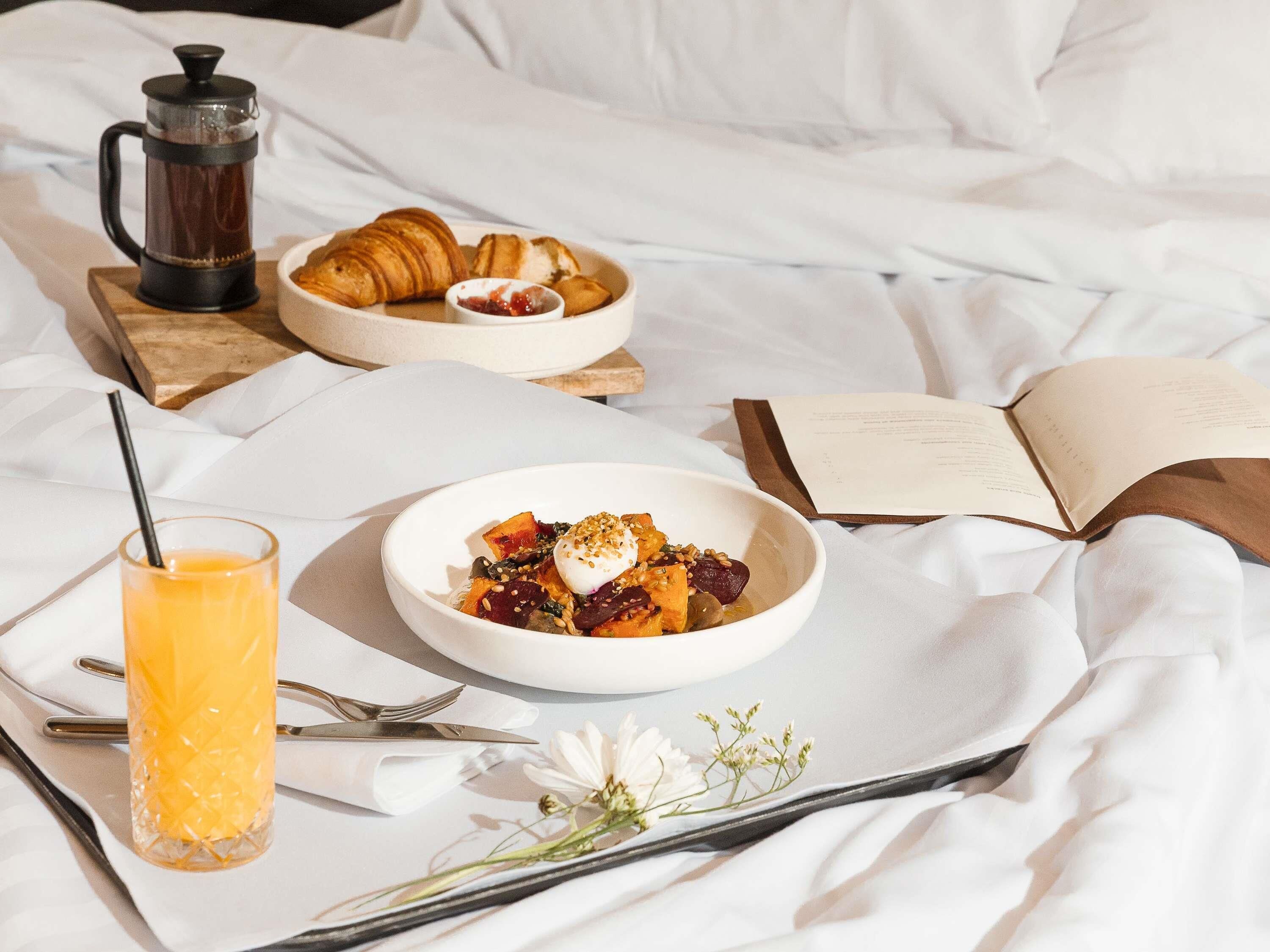 A breakfast setup with croissants, a coffee pot, a bowl of food, a glass of orange juice, and an open book on a bed.
