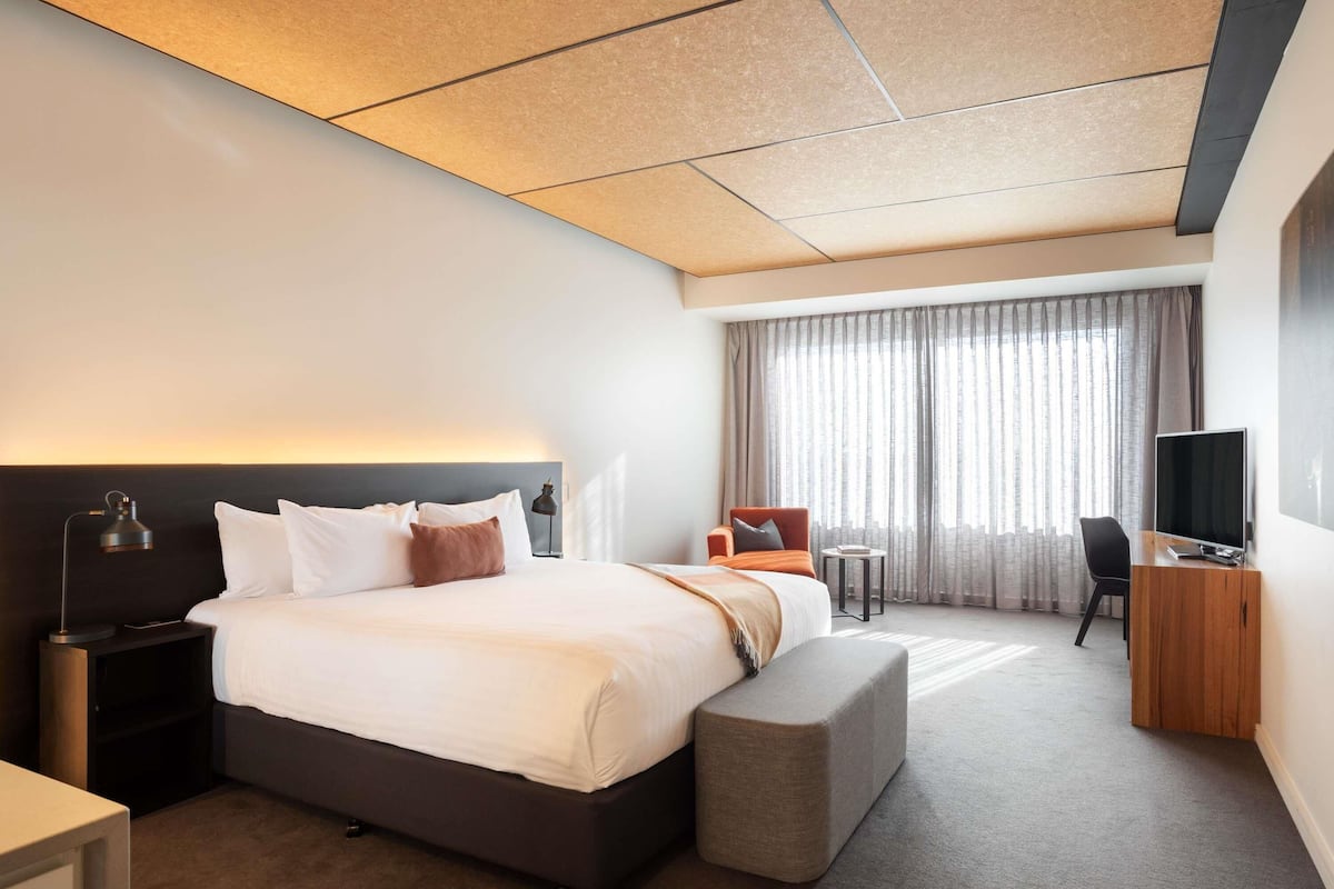A modern hotel room with a large bed, bedside tables, a desk with a chair, a TV, and a view of the city through large windows.