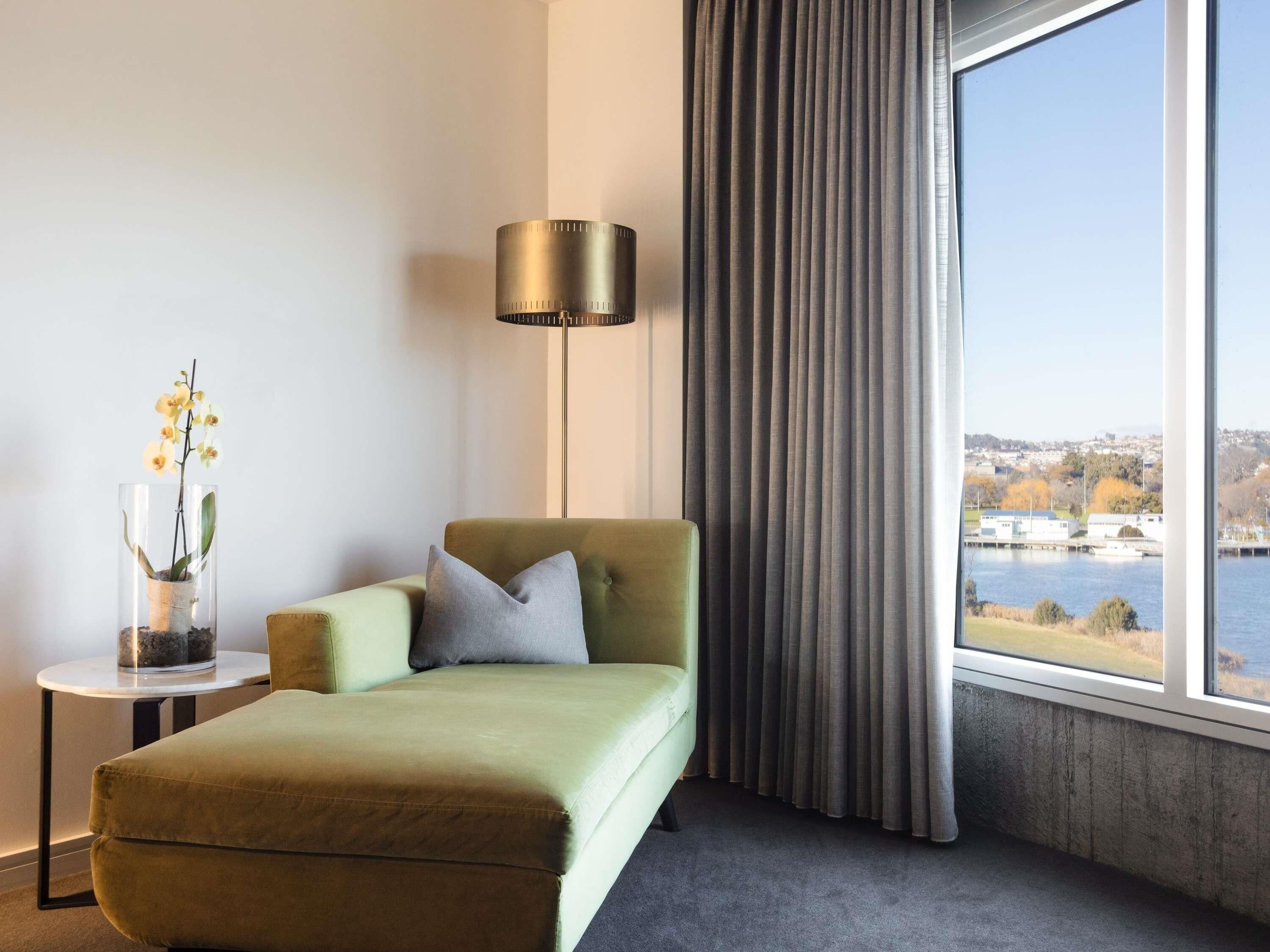 A modern hotel room with a green sofa, a small side table with a vase of flowers, a floor lamp, and a large window with a view of a body of water and buildings.