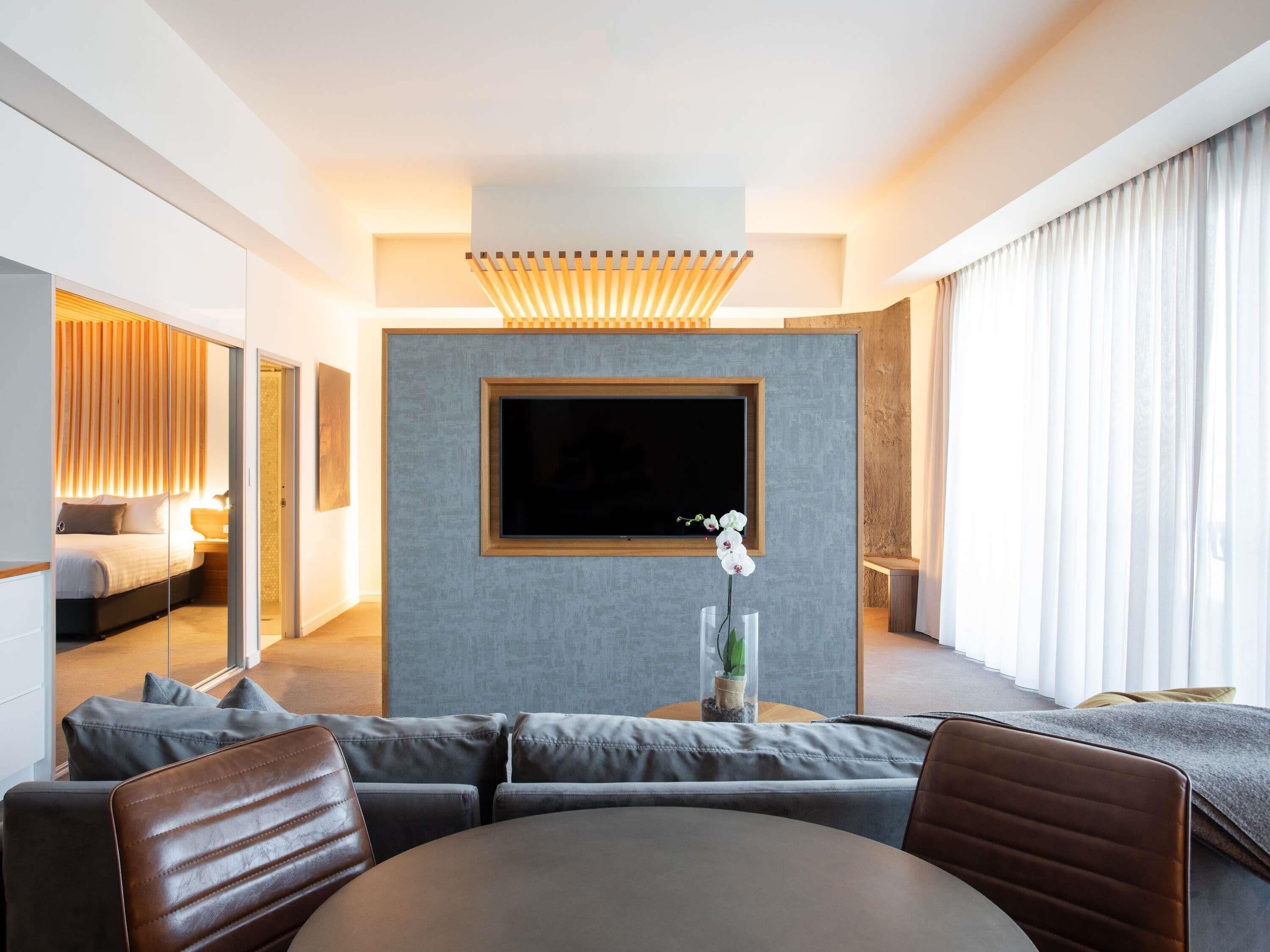 Ein modernes Hotelzimmer mit einem großen Flachbildfernseher, einer Couch, einem Esstisch und einem Spiegel.