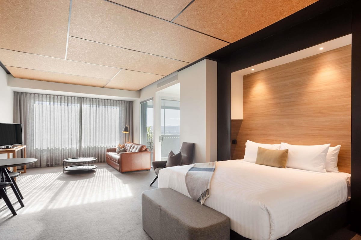 A modern hotel room with a large bed, a seating area with a sofa and armchairs, a TV, and a large window with curtains.