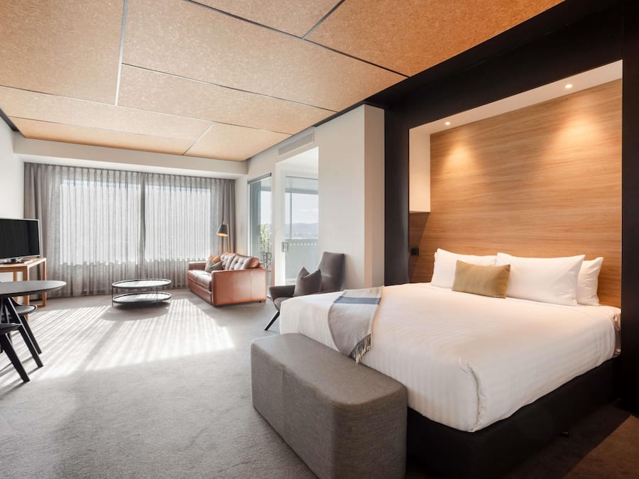 Ein modernes Hotelzimmer mit einem großen Bett, einer Sitzecke mit Sofa und Sesseln, einem Fernseher und einem großen Fenster mit Vorhängen.