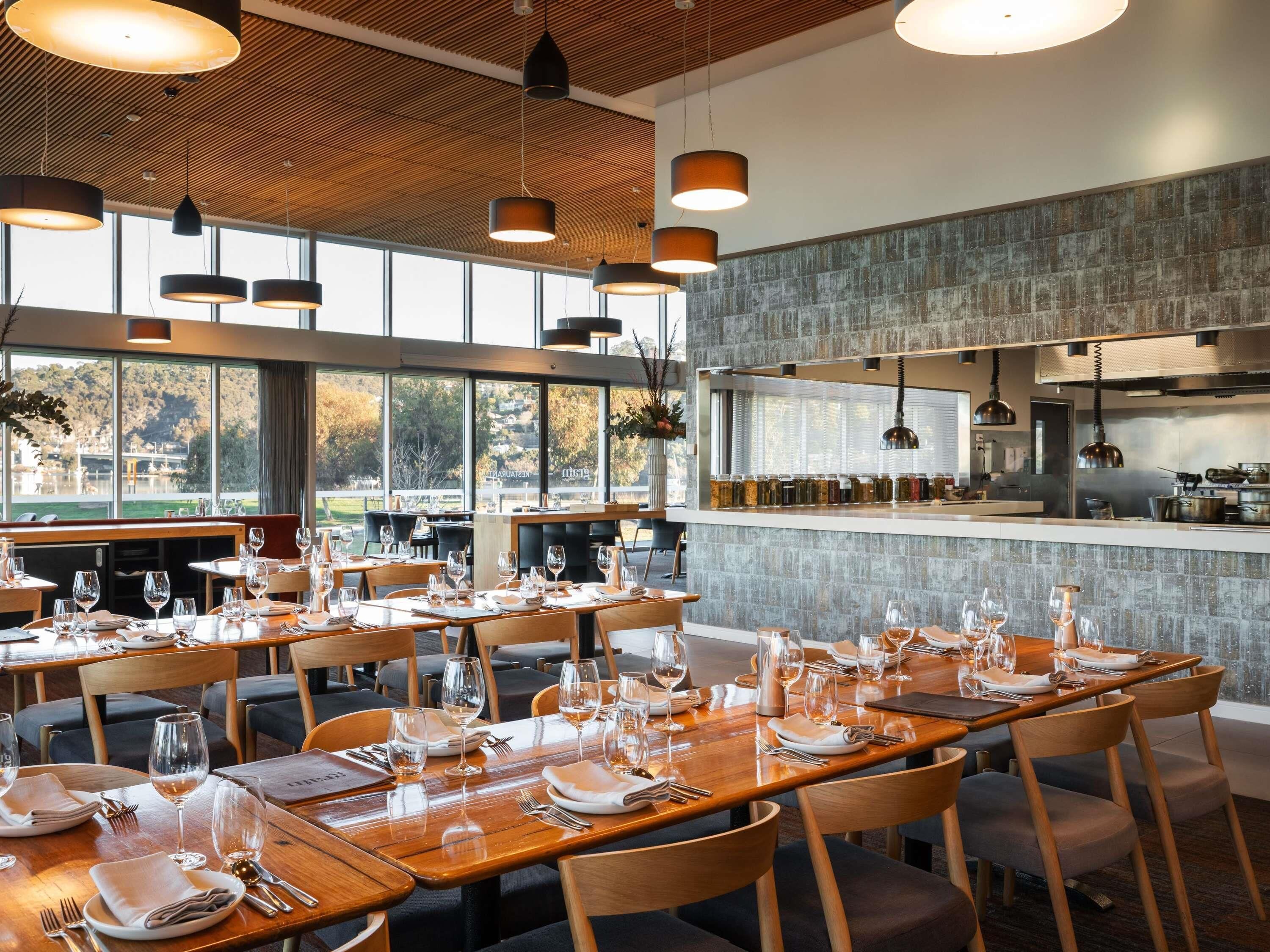 Un restaurante con mesas y sillas de madera, ventanas amplias y lámparas colgantes.