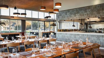 Un restaurante con mesas y sillas de madera, ventanas amplias y lámparas colgantes.
