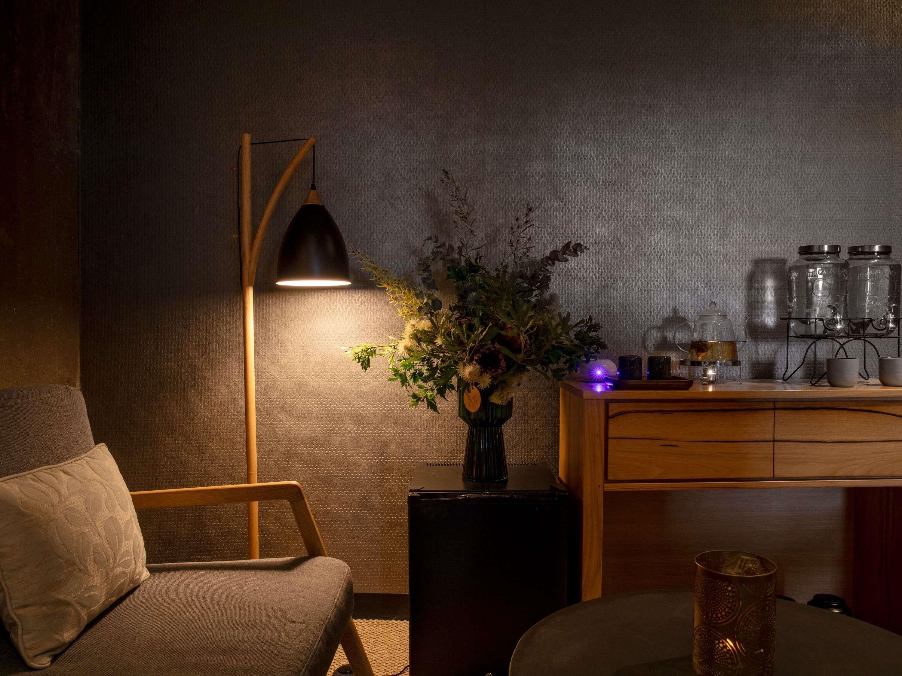 A cozy room with a wooden chair, a floor lamp, a vase with flowers, a coffee maker, and a small table with jars.