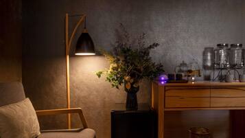 A cozy room with a wooden chair, a floor lamp, a vase with flowers, a coffee maker, and a small table with jars.