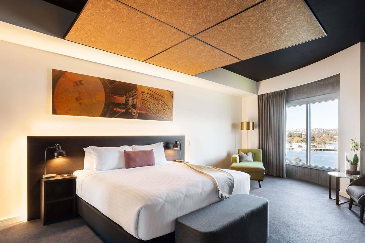 A modern hotel room with a large bed, a seating area, a view of the water, and a ceiling with a geometric design.