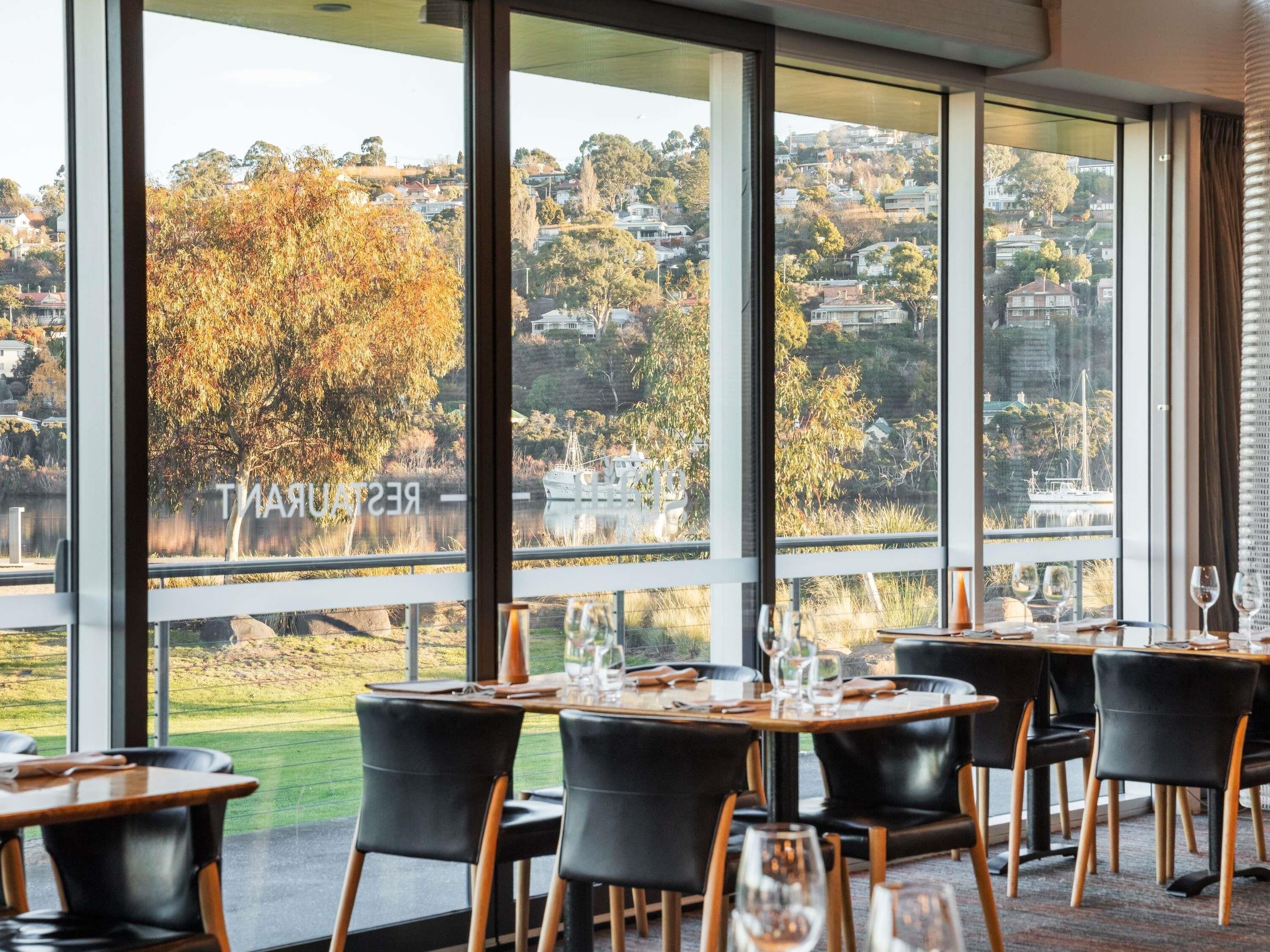 A restaurant with large windows offering a view of a hillside and a body of water. Tables are set with wine glasses and plates.