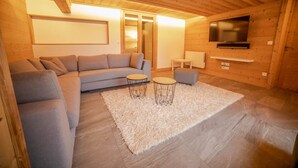 A cozy living room with a grey sectional sofa, two small round tables, a fluffy white rug, and a TV mounted on the wall.
