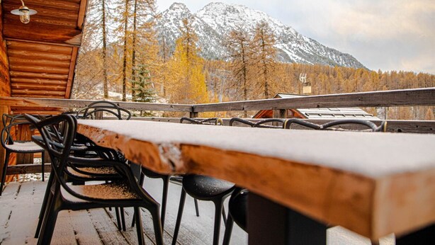 Una terraza de madera con una mesa y sillas, con vistas a una montaña nevada y árboles otoñales.