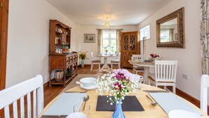Un coin repas avec une table en bois dressée pour deux personnes, entourée de chaises blanches. Un vase bleu rempli de fleurs est posé sur la table.