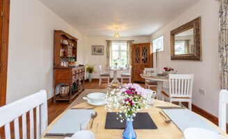 A dining area with a wooden table set for two, surrounded by white chairs. A blue vase with flowers is on the table.