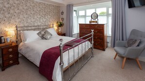 Une chambre à coucher avec un lit à baldaquin, une commode en bois, un fauteuil gris et un téléviseur fixé au mur.
