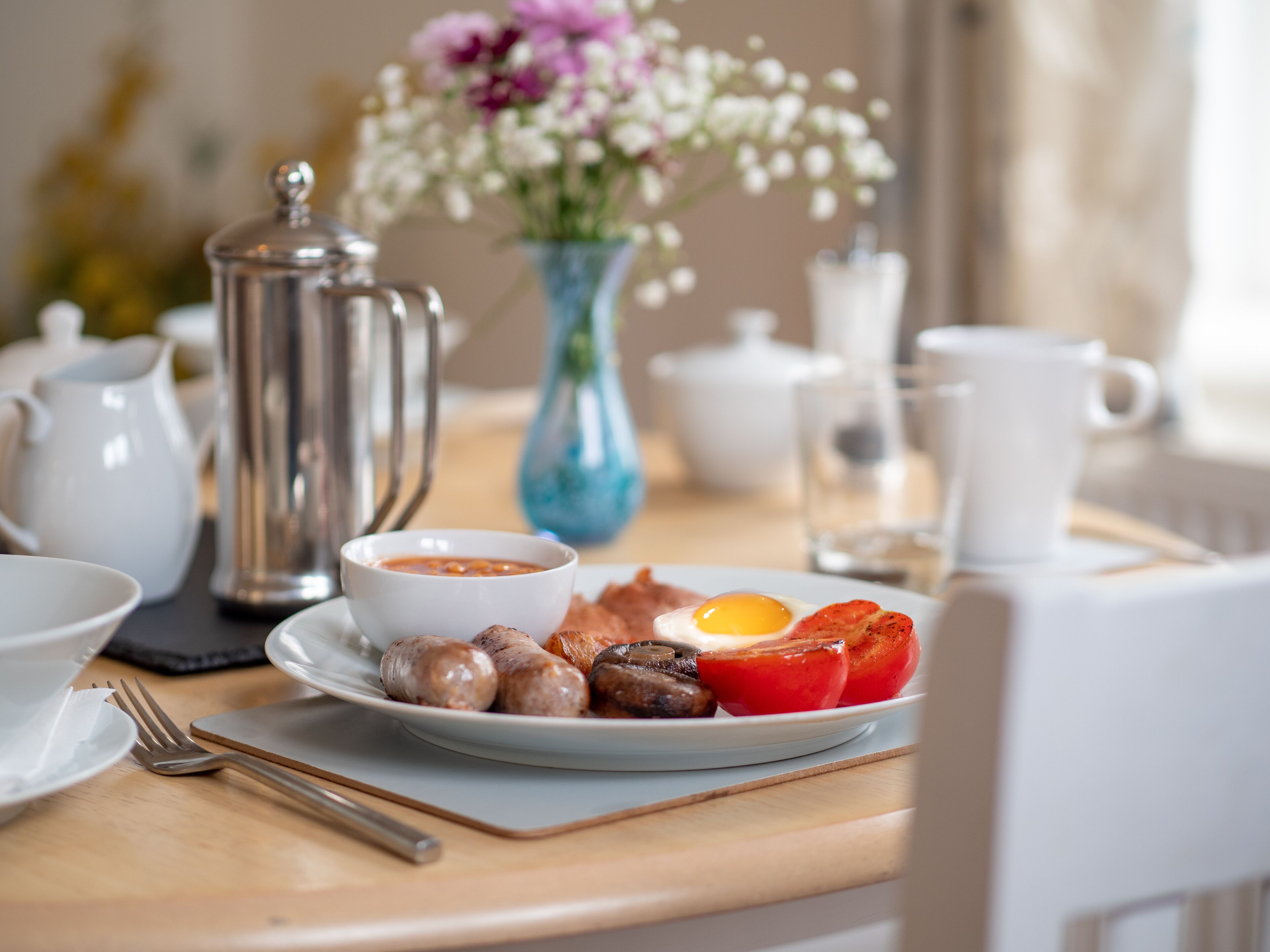Una colazione con un piatto di uova, salsiccia e pomodori, una caffettiera e un vaso di fiori.