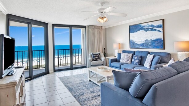 A modern living room with a large sofa, a flat-screen TV, and a balcony with a view of the ocean. - Phoenix VII 803 | Spacious, beach front, clean, comfortable! (Orange Beachg)