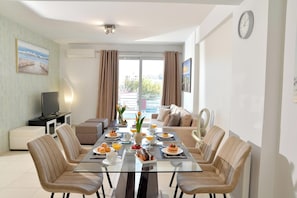 A modern living room with a glass dining table, beige sofas, a flat-screen TV, and a balcony with curtains. - Alekos Apartments 1 (Athens)