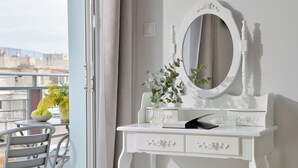 A white vanity with a round mirror, a vase with green foliage, and a small table with a bowl of fruit. - Alekos Apartments 1 (Athens)