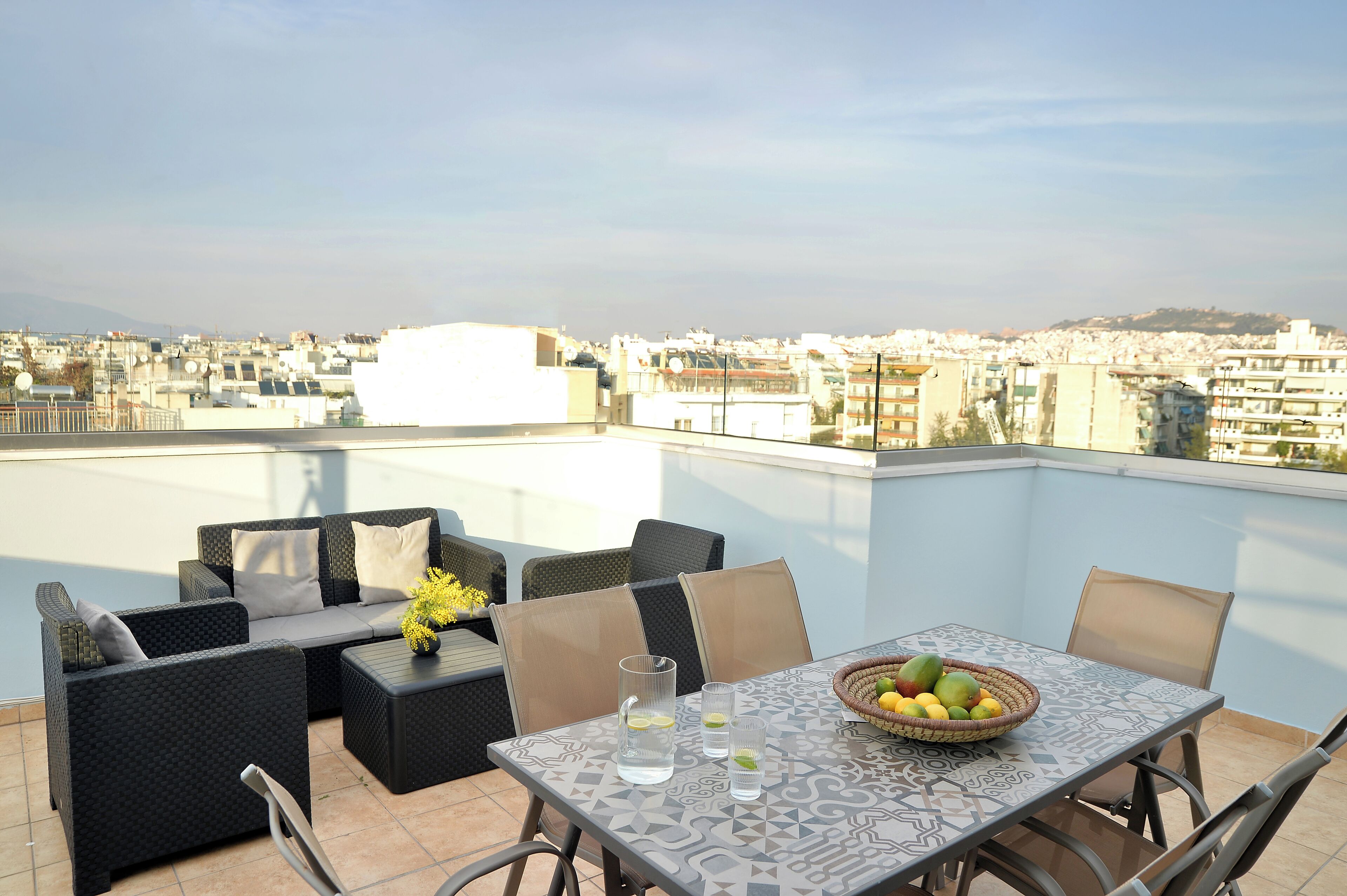 A rooftop terrace with outdoor furniture, a table with a fruit bowl, and a city view.