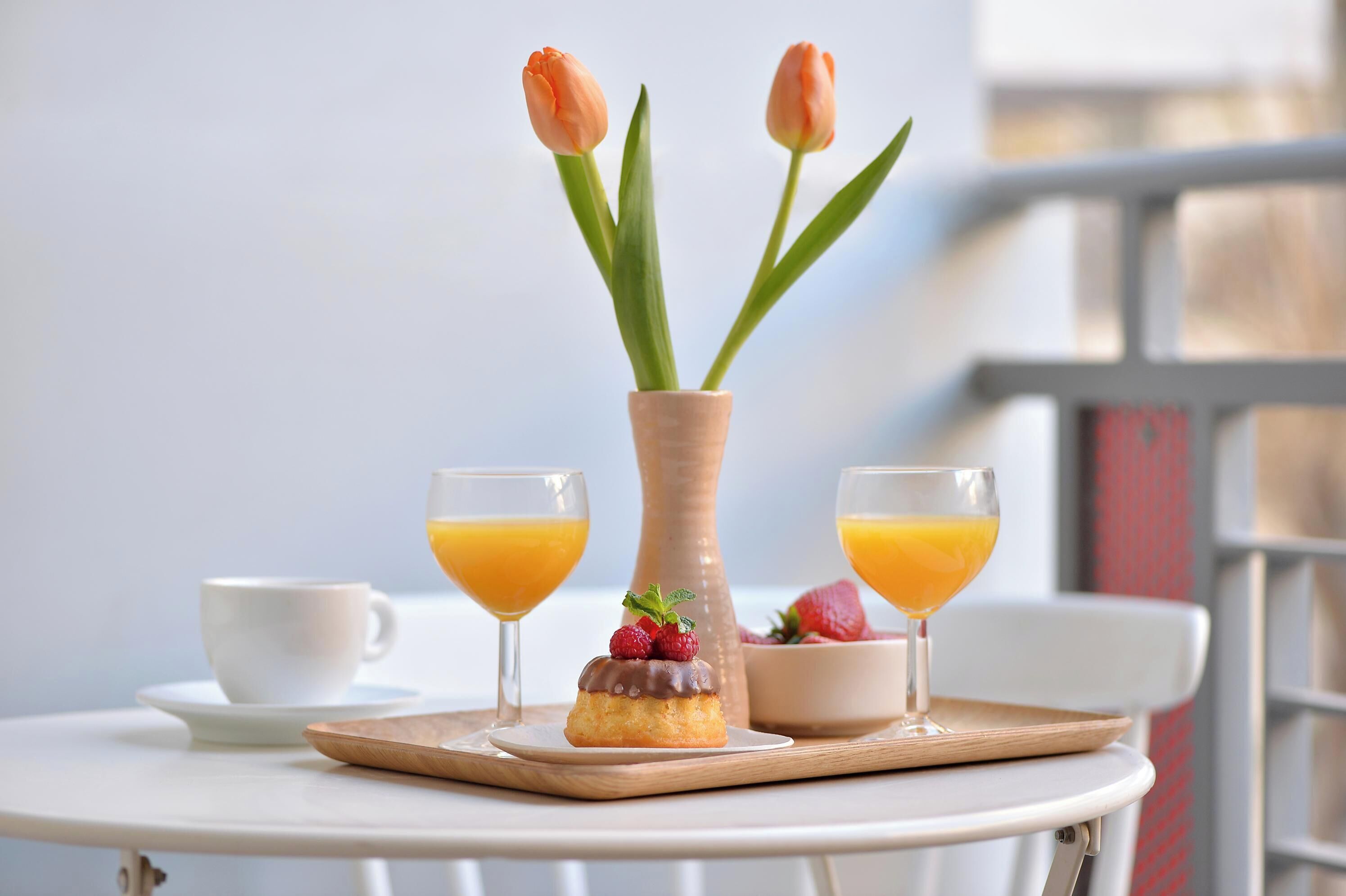 Een ontbijtsetting met een witte tafel, twee glazen sinaasappelsap, een witte kop, een klein houten dienblad met een gebakje en een vaas met twee oranje tulpen.