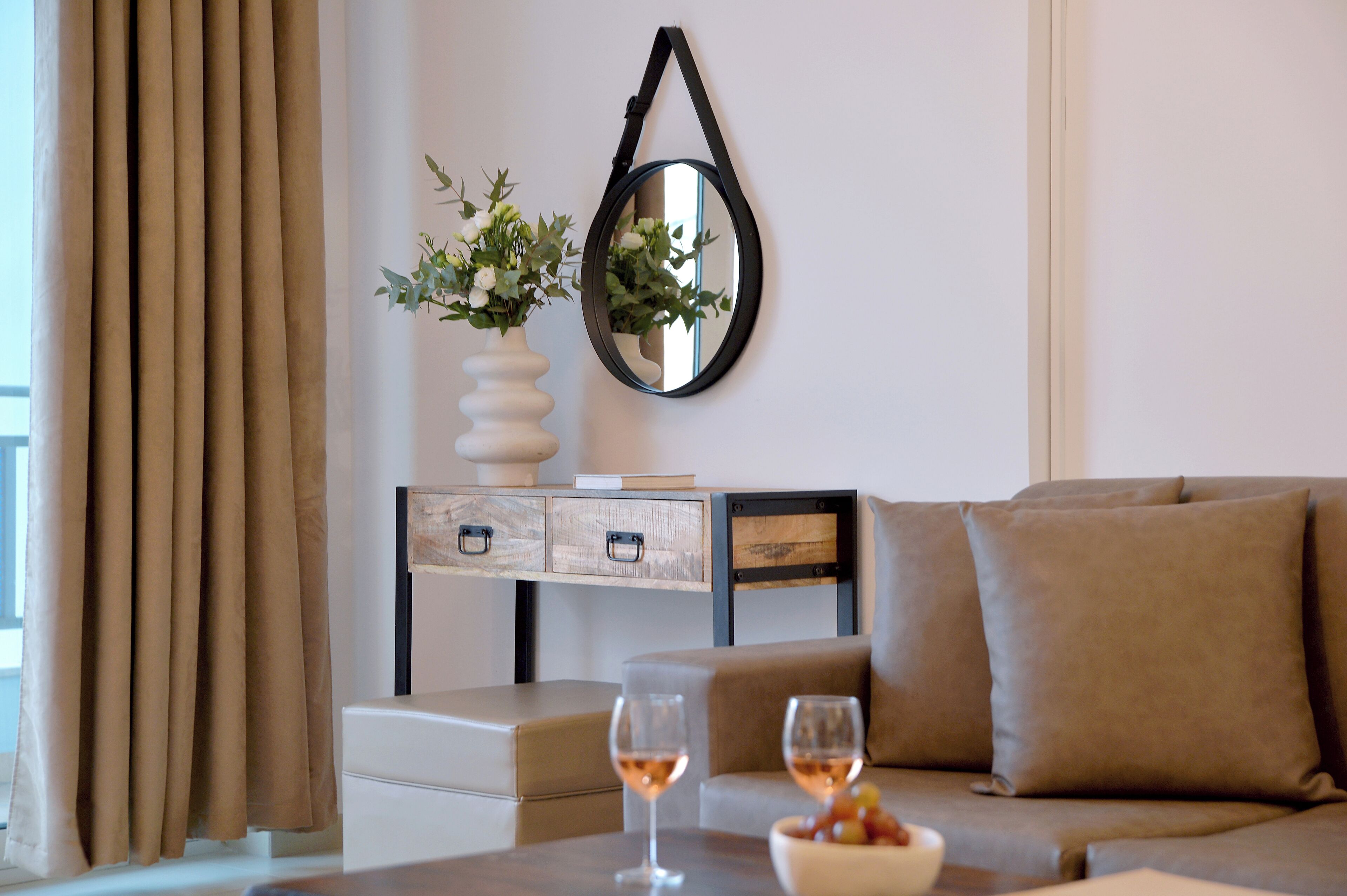 A modern living room with a sofa, a coffee table with two glasses of wine, a vase with flowers, and a wall-mounted mirror.