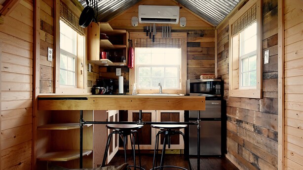 A compact kitchen with wooden walls, a bar stool, a sink, and a microwave.