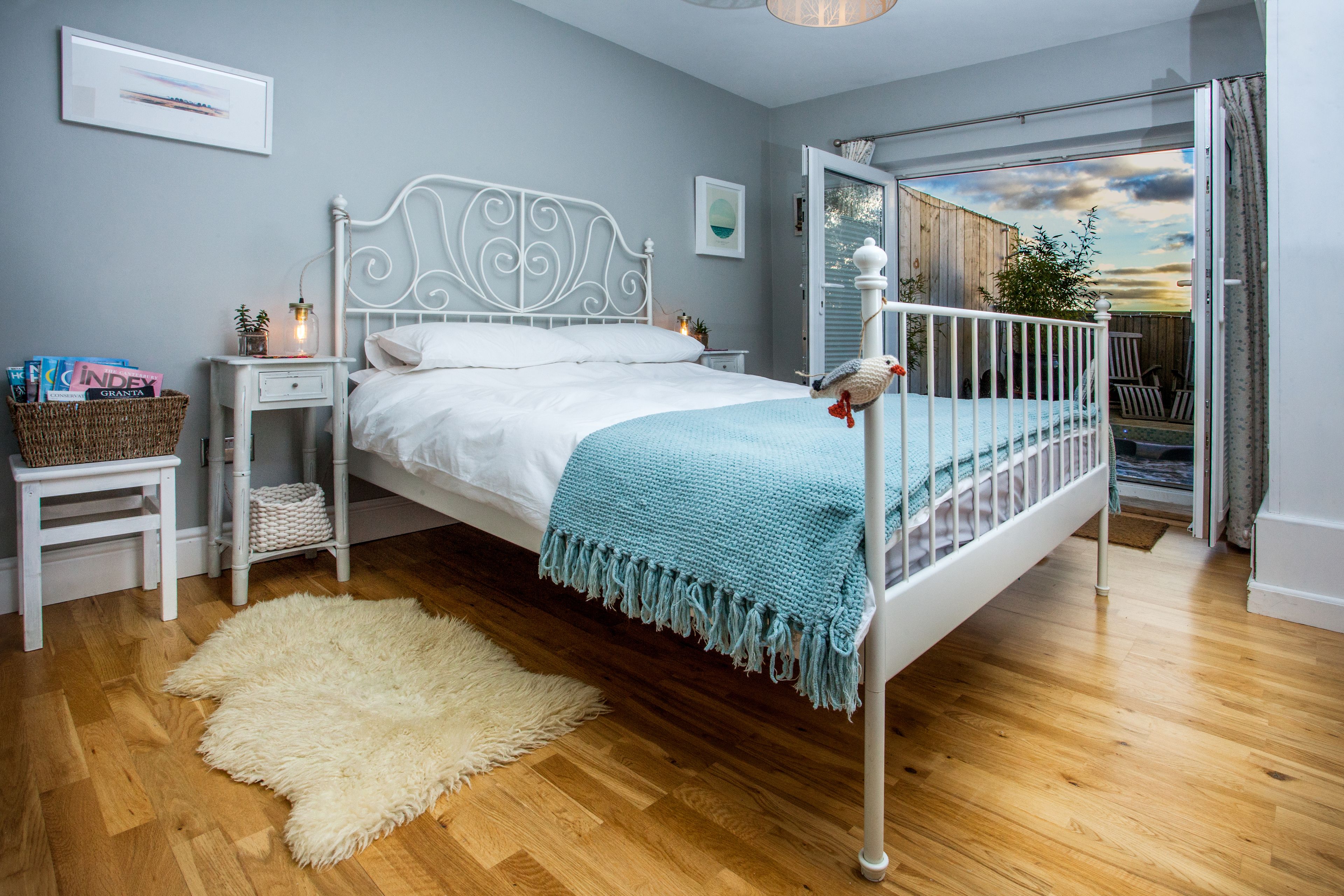 A bedroom with a white metal bed frame, a blue throw blanket, a white fluffy rug, and a view of the outdoors through an open door.