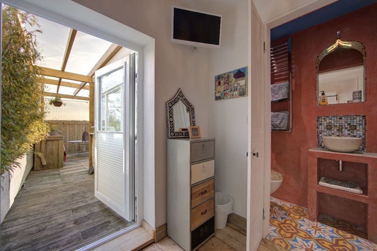 Quarto com deck de madeira, porta de vidro deslizante para o exterior, penteadeira, espelho, painel de azulejos e televisão fixada na parede.