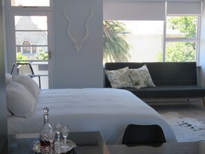 A bedroom with a bed, a table with a bottle and glasses, a sofa with pillows, and a view of greenery outside.