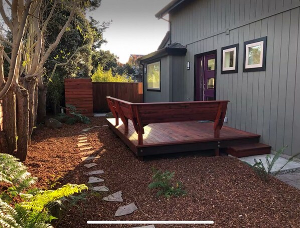Une terrasse en bois avec un banc, entourée d’un jardin agrémenté de paillis et de pierres de pas, menant à une maison à l’extérieur gris et à la porte violette.