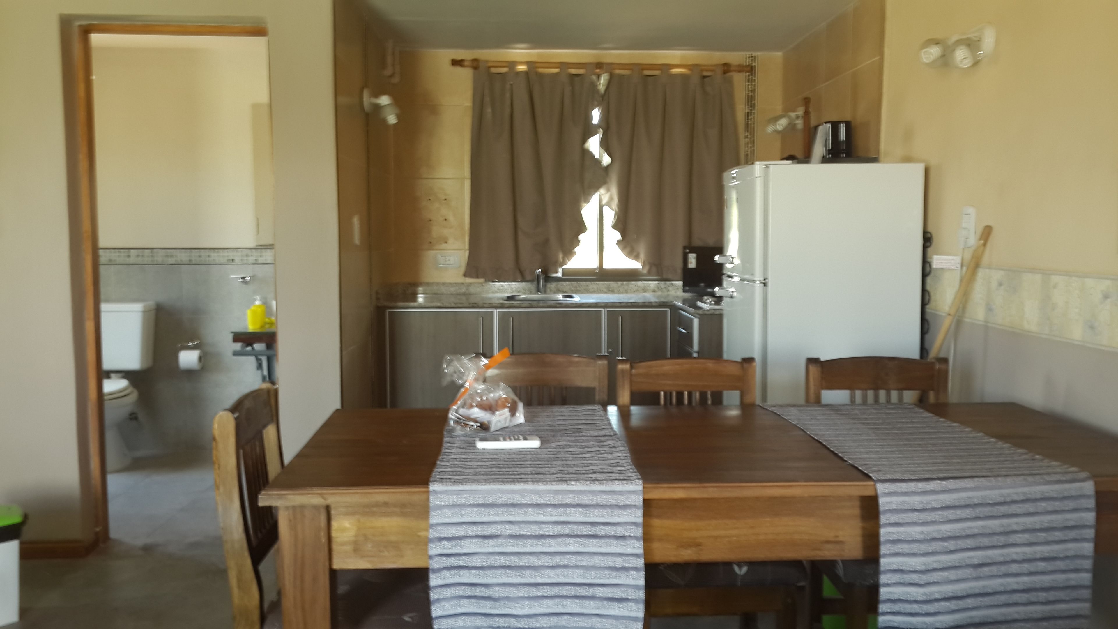 Eine kleine Küche mit Kühlschrank, Spüle und Grundausstattung. Im Vordergrund steht ein Esstisch mit Stühlen, im Hintergrund ist ein Fenster mit Vorhängen zu sehen.