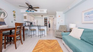A modern living room with a teal sofa, wooden dining table, and chairs. A kitchen is visible in the background with white cabinets and a ceiling fan.