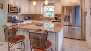 A kitchen with white cabinets, a stainless steel refrigerator, a microwave, and a large island with two wicker chairs.