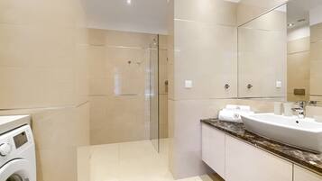 Un baño moderno con ducha envuelta en vidrio, lavadora integrada y un lavamanos grande con encimera de mármol.