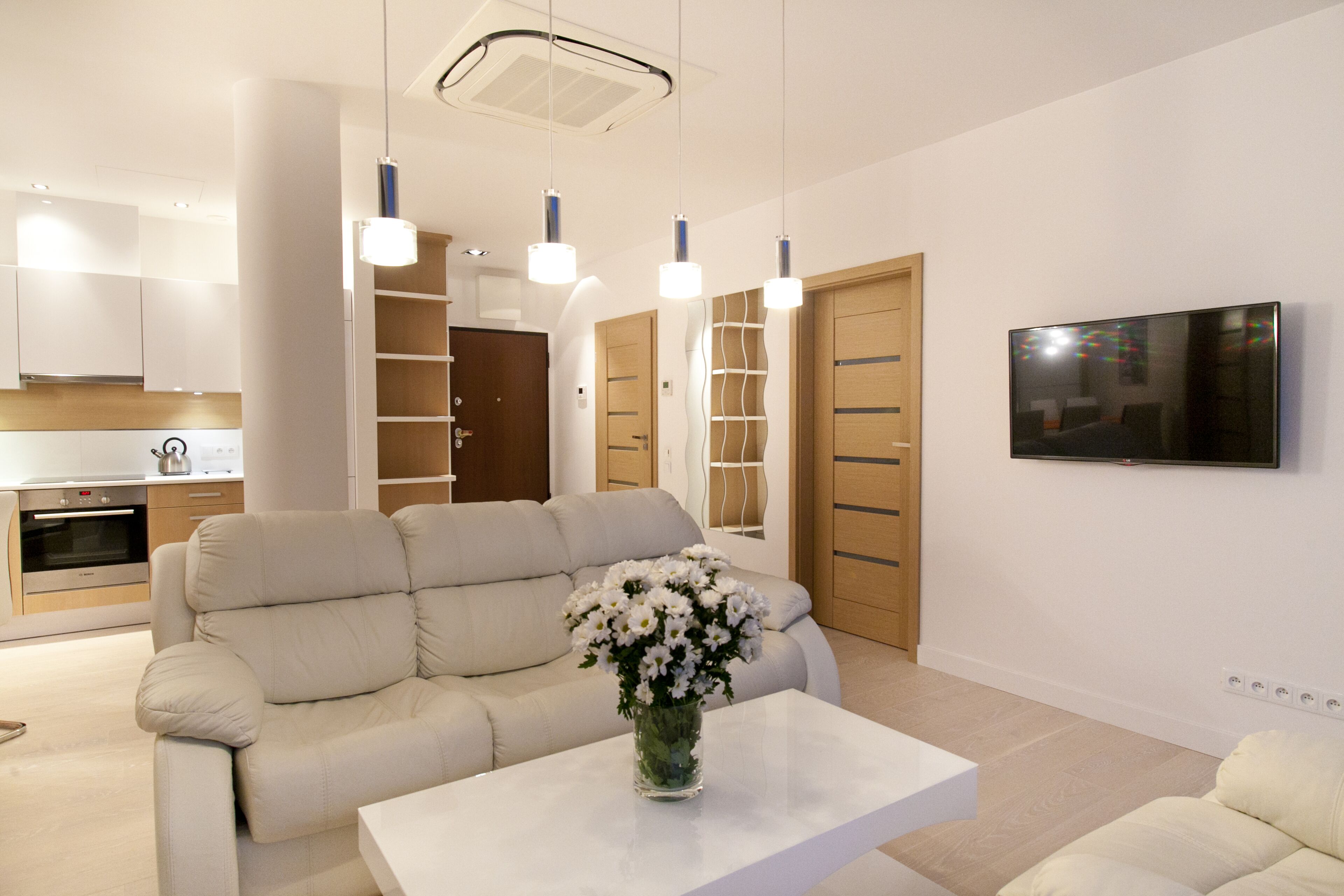 Una sala de estar moderna con un sofá beige, una mesa de centro blanca y un jarrón con flores. Hay una zona de cocina con armarios y un horno empotrado. Un televisor de pantalla plana está montado en la pared.