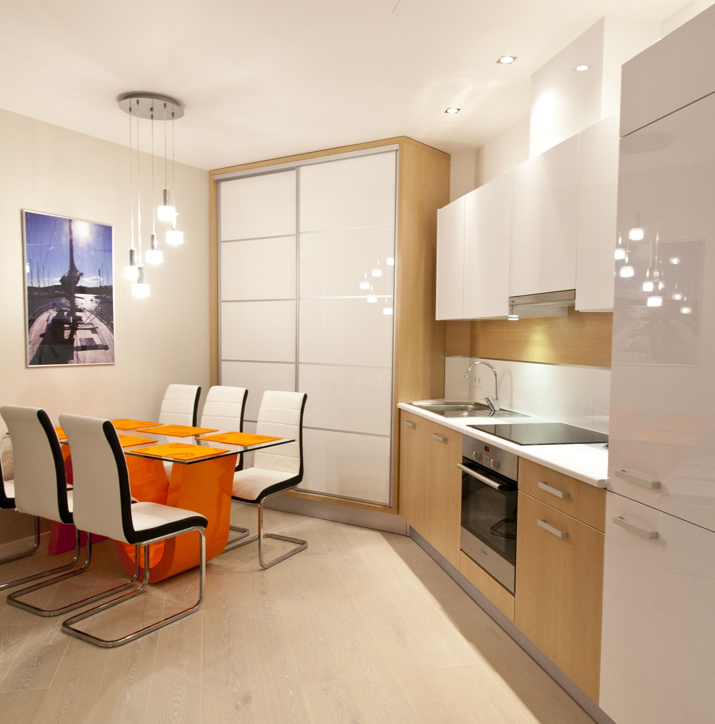Een moderne keuken met witte kasten, een roestvrijstalen spoelbak en een ingebouwde oven. Er is een eethoek met een glazen tafel en stoelen.
