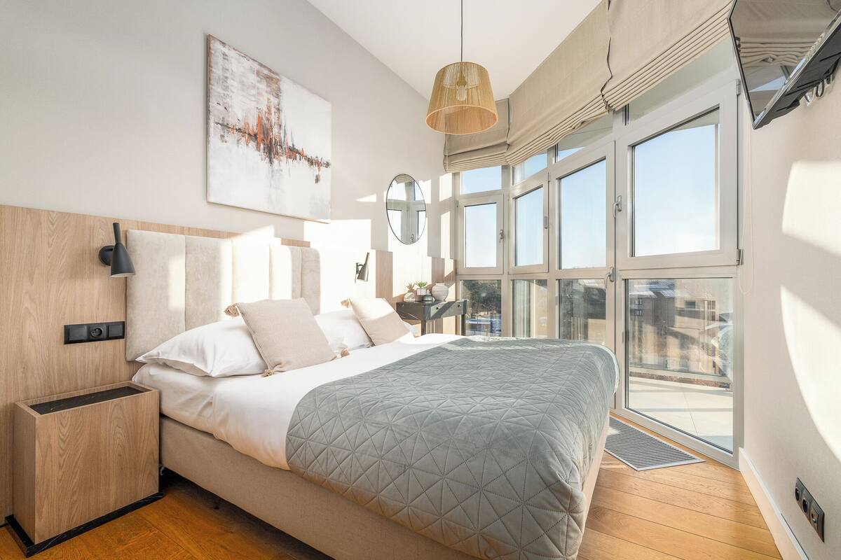 Une chambre moderne avec un grand lit, une table de chevet, une lampe fixée au mur et une grande fenêtre donnant sur l’extérieur.