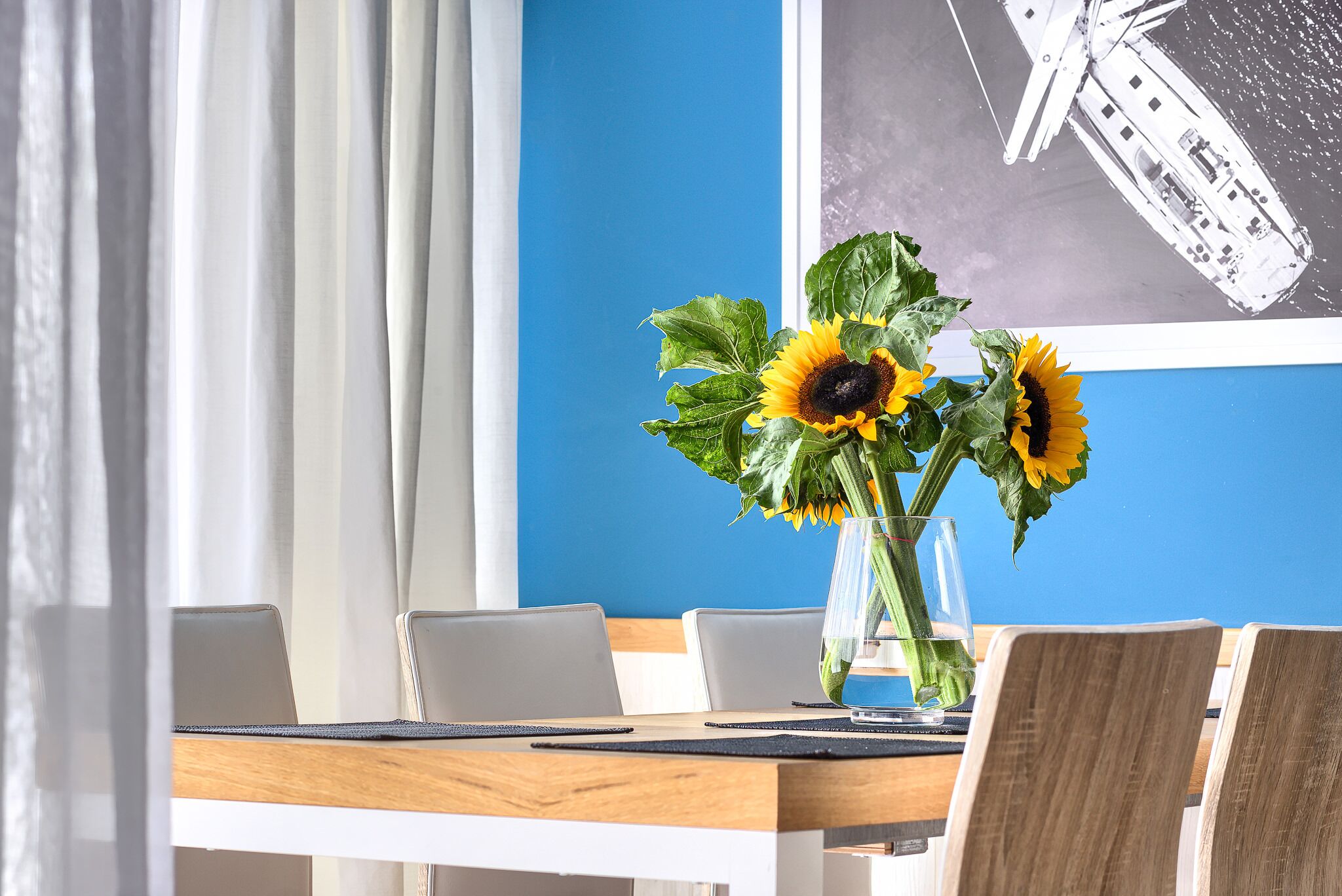 Un coin repas avec une table en bois, des chaises blanches et un vase rempli de tournesols.
