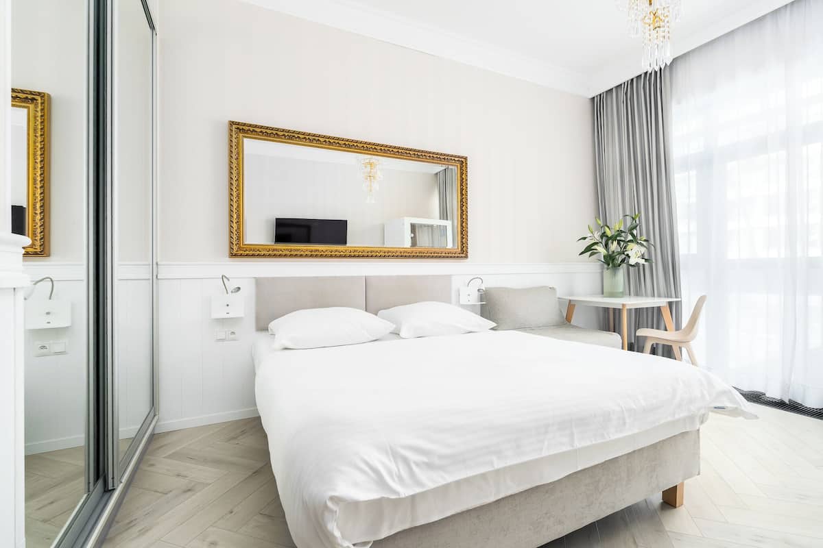 Une chambre d’hôtel moderne avec un grand lit, une table de chevet, une chaise, une petite table et un grand miroir avec un cadre doré.
