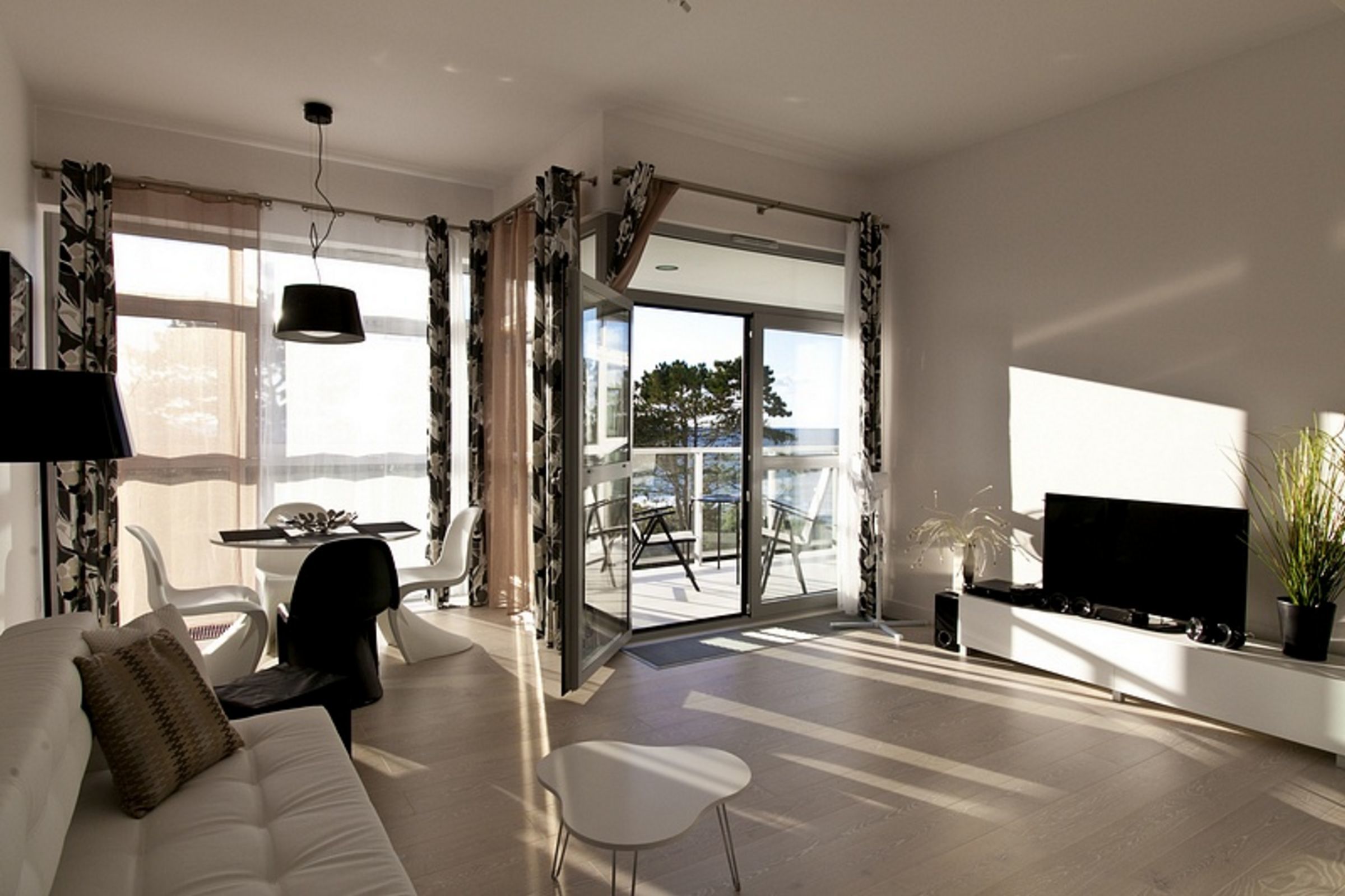 A modern living room with a sofa, a dining table with chairs, a TV, and a balcony with a view.