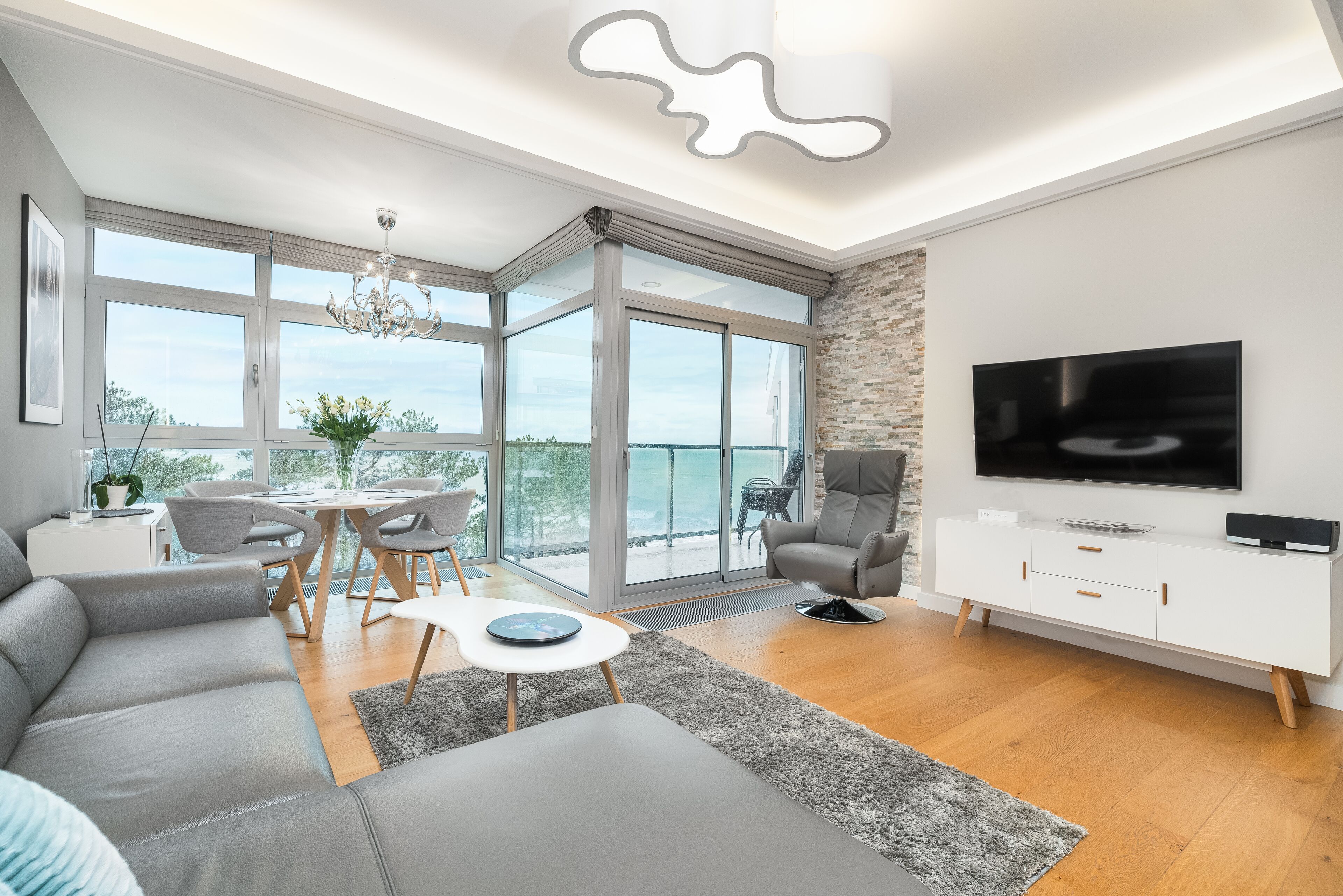 Een moderne woonkamer met een grote televisie, grijze banken, een eethoek met tafel en stoelen, en een balkon met uitzicht.
