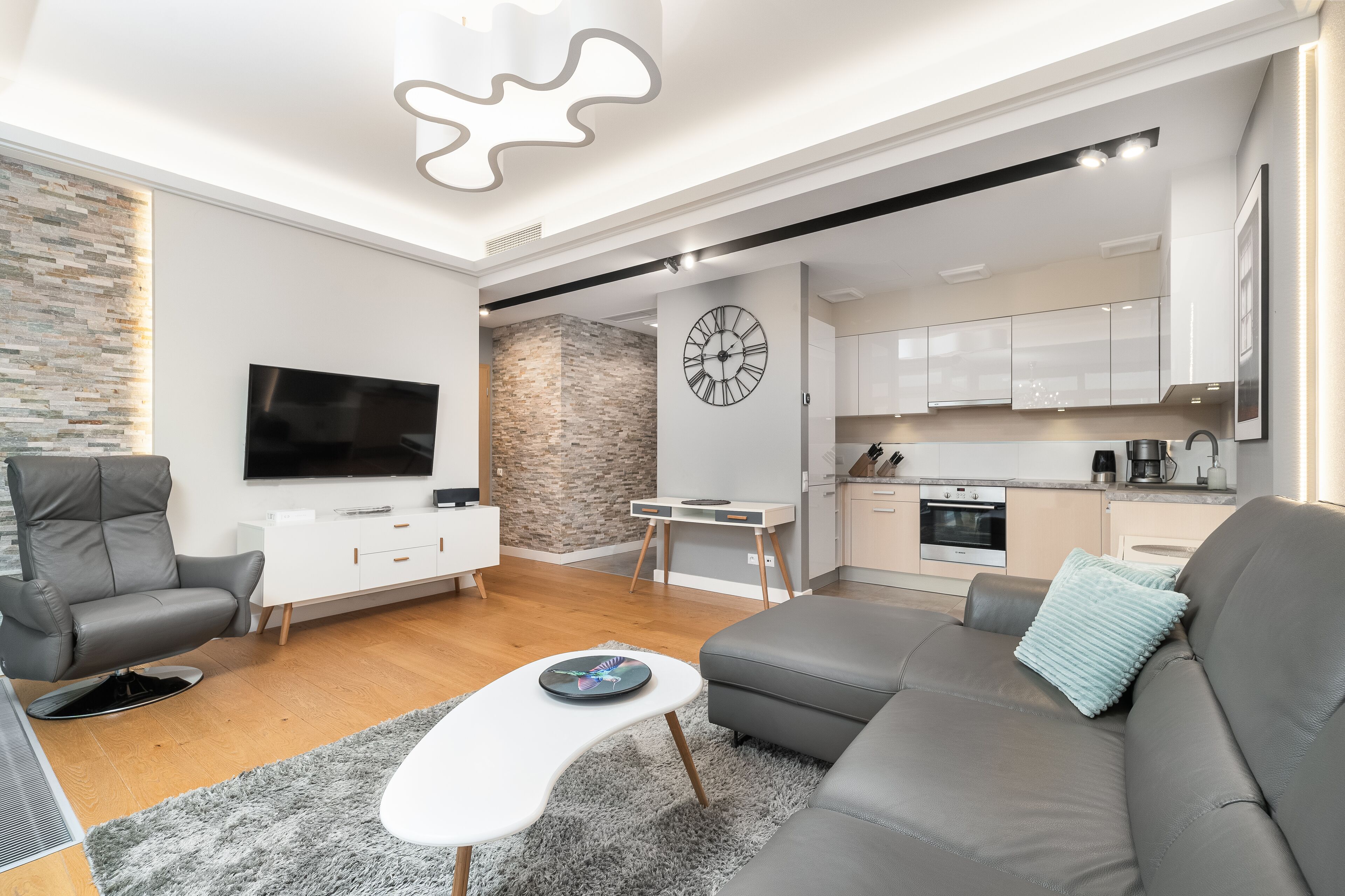 A modern living room with a grey sofa, a white coffee table, and a grey armchair. There is a flat-screen TV mounted on the wall, a clock, and a kitchen area visible through an open door.