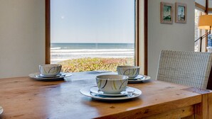 A wooden table with three bowls and saucers, a wicker chair, and a view of the ocean through large windows. - Ocean Front Home on a Sandy Beach Near Yachats! (Yachats)