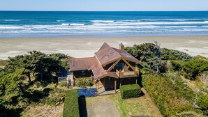 A coastal house with a large deck, surrounded by greenery and overlooking the ocean. - Ocean Front Home on a Sandy Beach Near Yachats! (Yachats)