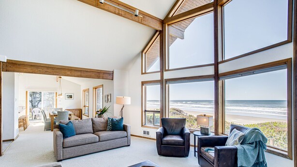 A spacious living room with large windows overlooking a beach, furnished with a grey sofa, armchairs, and a coffee table. - Ocean Front Home on a Sandy Beach Near Yachats! (Yachats)