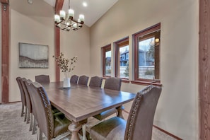 A dining room with a large wooden table, six upholstered chairs, a vase with flowers, a chandelier, and large windows with a view of a house.