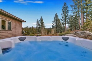 Un jacuzzi en un patio trasero con una casa de madera y altos pinos al fondo.